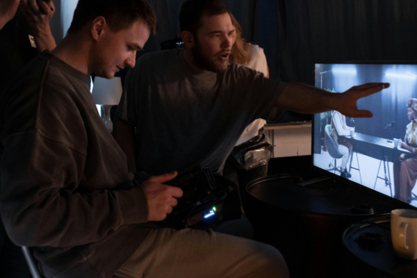 2 people sat in front of a large tv, one person looks to be directing a podcast involving 2 people.