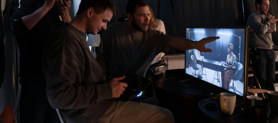 2 people sat in front of a large tv, one person looks to be directing a podcast involving 2 people.